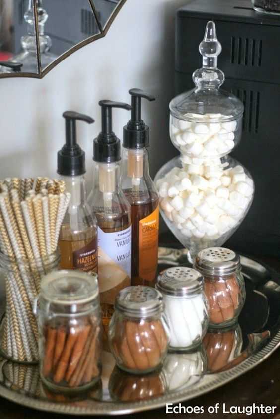 A tray on a countertop with supplies for a coffee station - syrup dispensers, shakers with cinnamon and sugar, mason jar with straws and a canister with marshmallows.