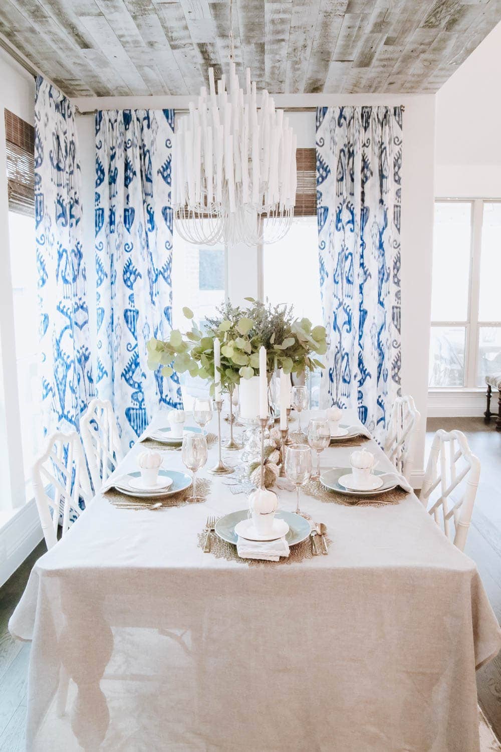 blue and white drapes, white pumpkins in a painted white tin, gold charger placemats with a white plate. Eucalyptus leaves in a tall vase in the center of the table and gold tapered candle stick holders.