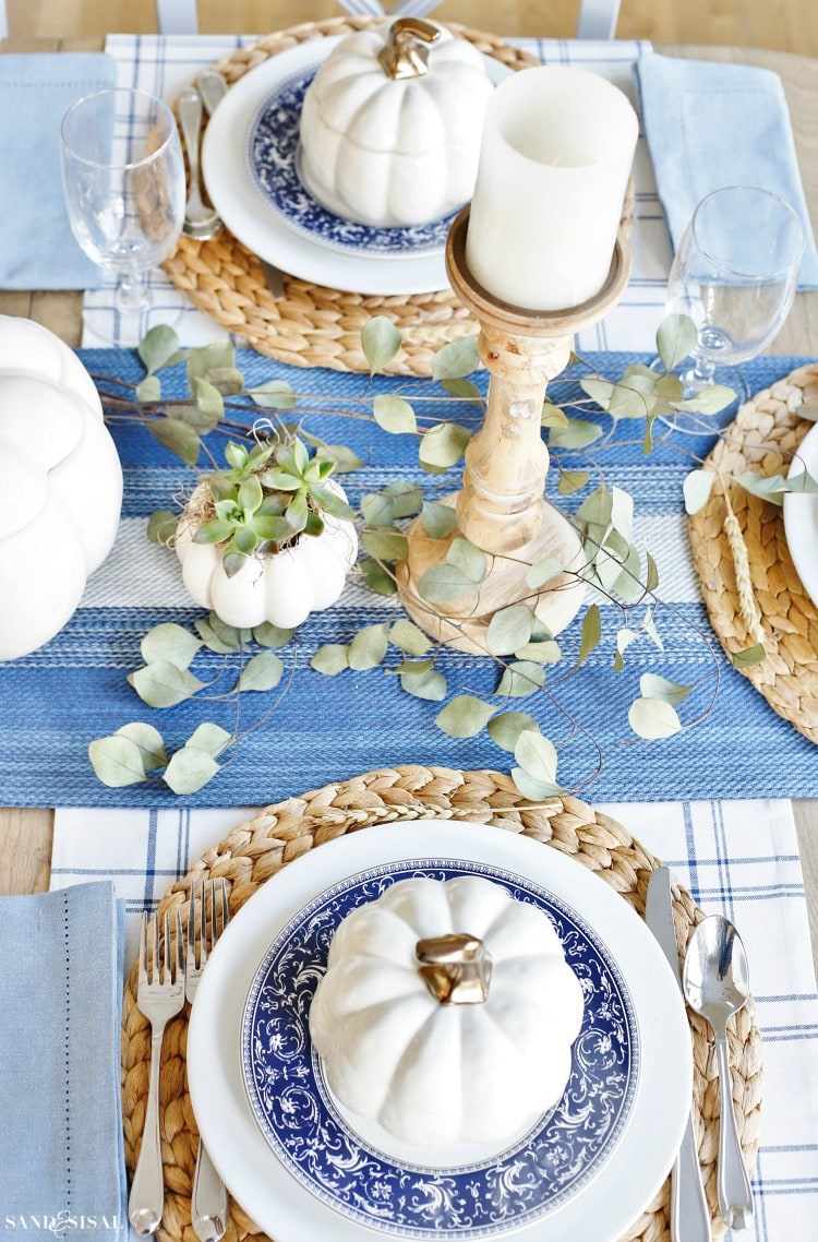 A classic blue and white Thanksgiving table setting featuring blue and white plates with a white pumpkin on top of the plate. Wood candlesticks and touches of eucalyptus leaves in the center of the table.