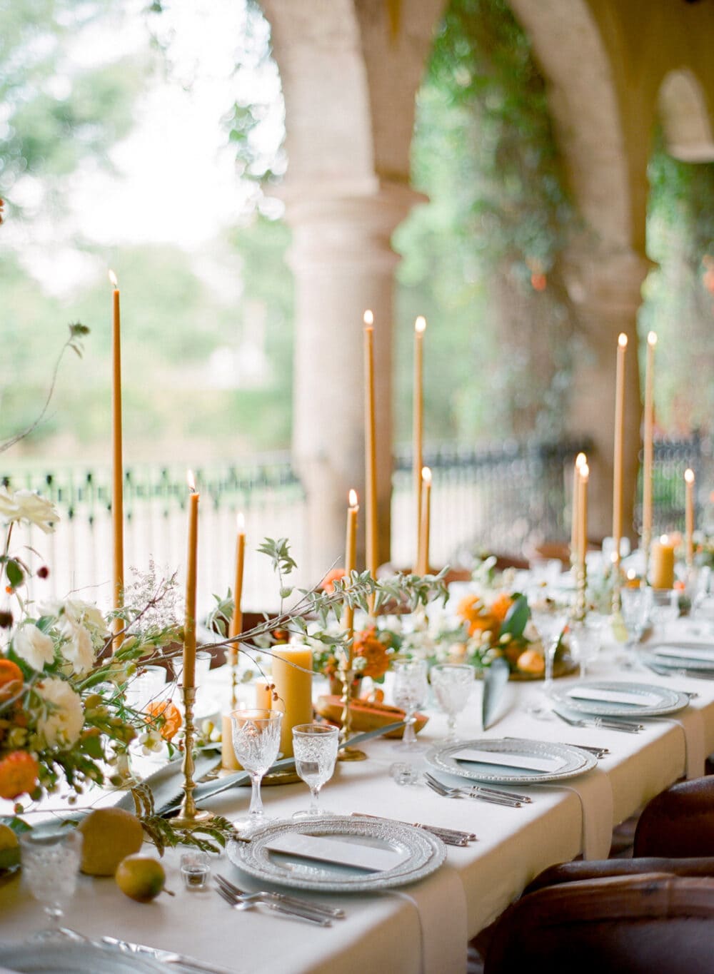 An outdoor Thanksgiving tablescape featuring soft amber colors. Tapered candles in a mustard color, groups of fall florals spread throughout the center of the table. Beautiful white layered dishes.