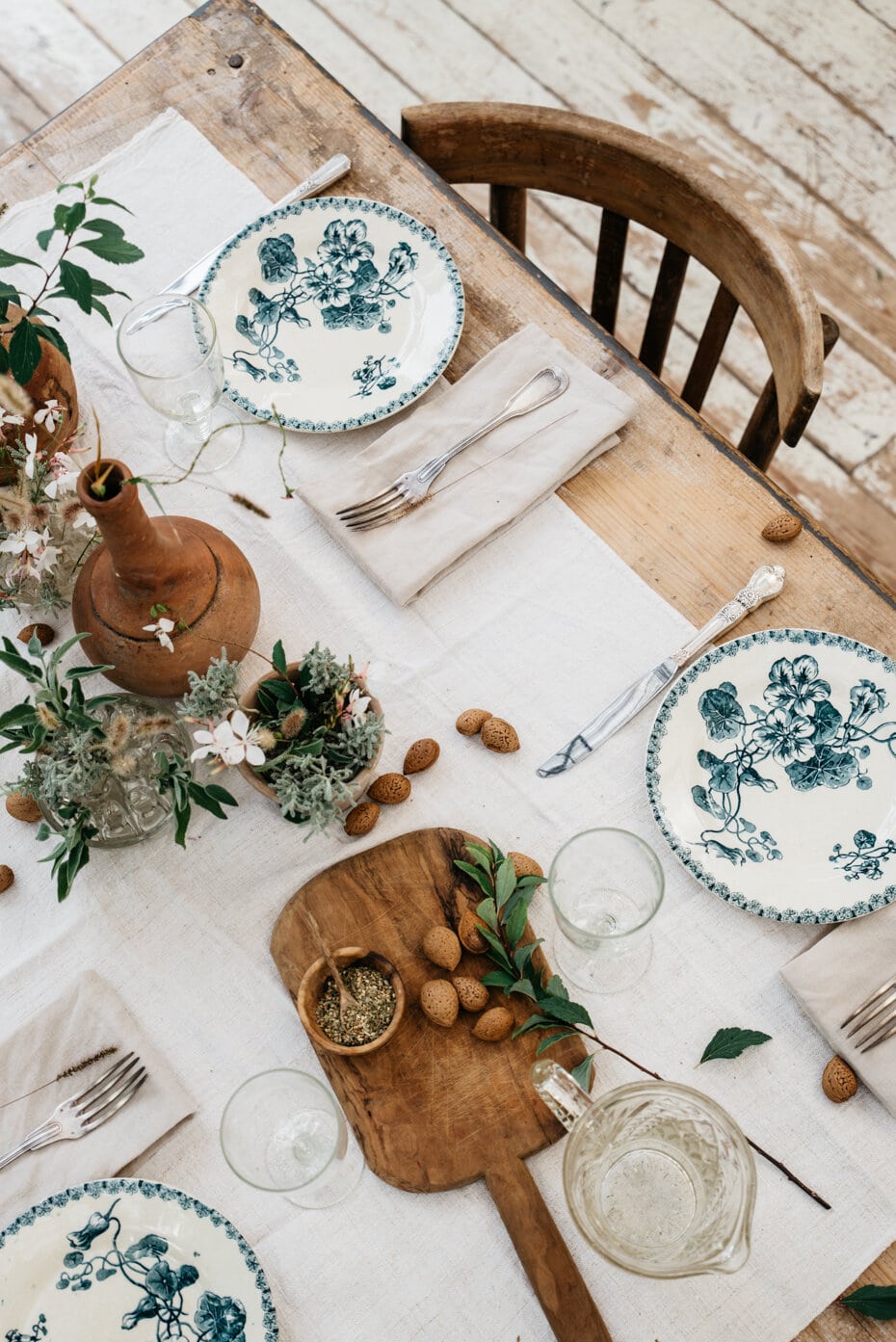 A rustic table with chippy white paint, blue and white vintage plates and beige linen napkins for place settings. VIntage cutting boards and wood candlesticks and eucalyptus leaves for the center of the table. 