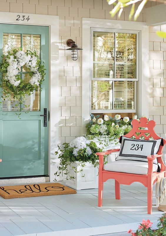 Love the color pops on this spring front porch! A light turquoise wooden door is decorated with a white and green floral wreath. A bright coral colored deck chair sits with white fabric throw pillows and curtains. The floral arrangements are a mix or white and green flowers. #spring #springporch #springdecorating #springfrontporch