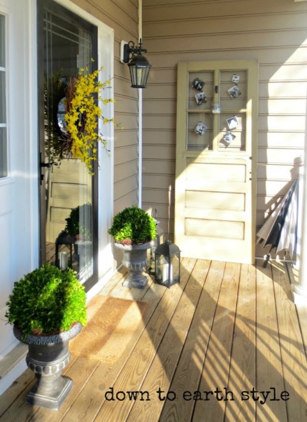 Spring front porch with stone planters filled with green shrubbery. A weathered wood door decorated with vintage photographs leans against the wall. On the door, there is a twig wreath with bright yellow flowers. Gray metal lanterns flank the planters.