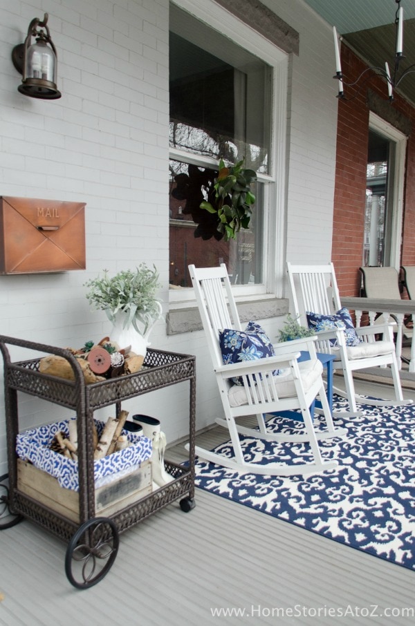 This spring front porch is decorated with a blue theme. White rocking chairs have blue floral patterned throw pillows. A vintage tea cart holds small planters, rain boots, and spring decor. A white and blue oriental pattern rug sits in the center of the porch. 