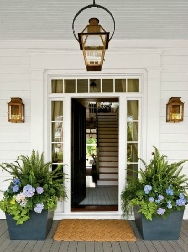 An open front door is flanked by two large square stone planters filled with tropical plants. Purple and blue flowers accent the green leaves. 