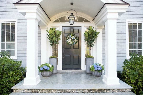 This spring themed front porch is decorated with large planters filled with spring floral arrangements. Two planters flank the door holding fig leaf plants. A white, blue, and green wreath adorns the front door. This color scheme is matched in the smaller, round planters on either side of the porch. 