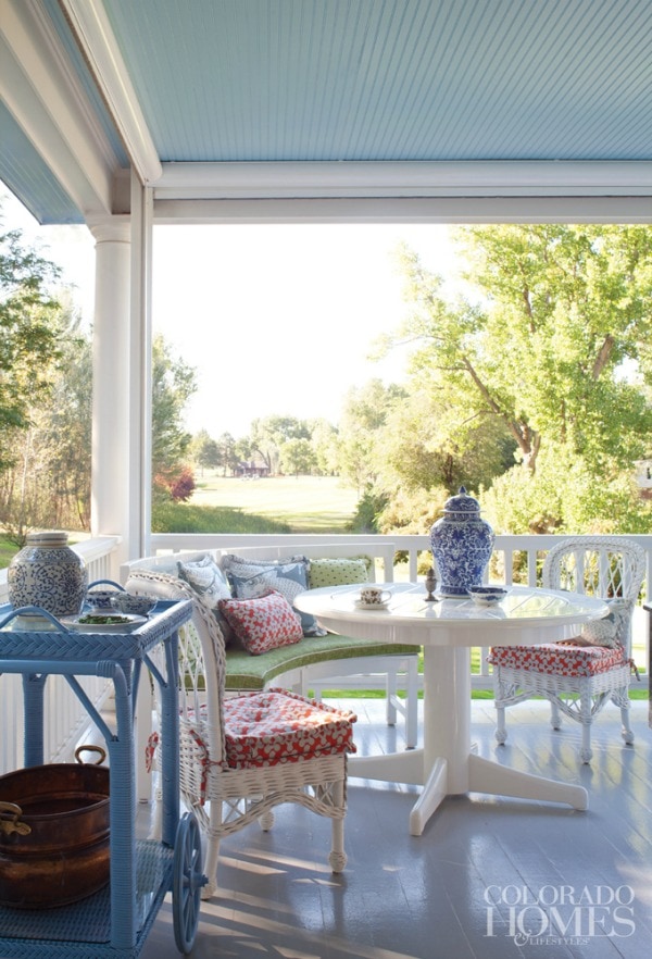 This spring front porch is decorated with beautiful white furniture. The cushions on the chairs are a red, blue, and white floral fabric, accented with pops of green. A periwinkle blue tea cart holds ceramic planters and a vintage washing tub. There is a blue oriental patterned urn on the table. 