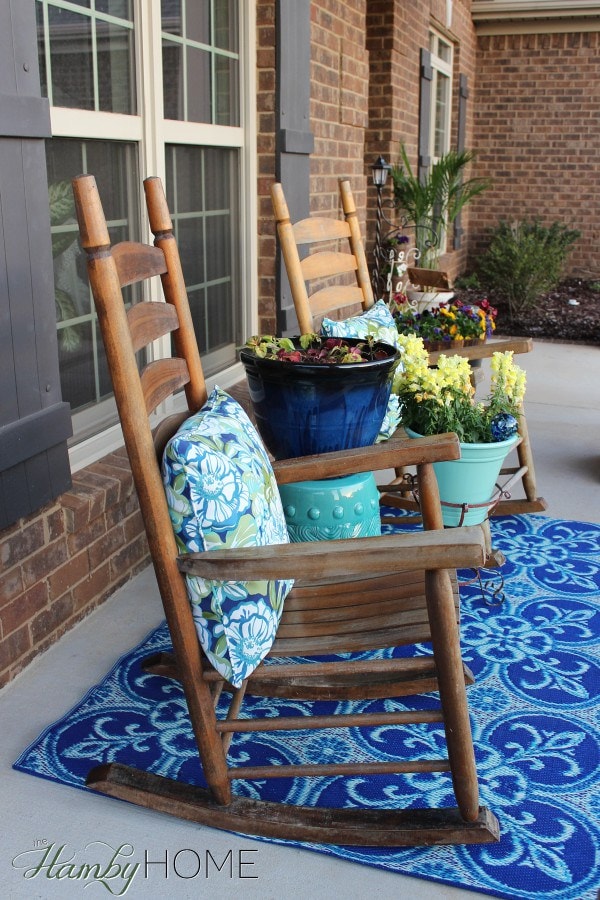 This spring front porch is packed with bright blues. The dark wooden rocking chairs have blue floral fabric throw pillows. Dark and light blue planters hold blooming flowers. A blue fleur-de-lis rug sits in the center.