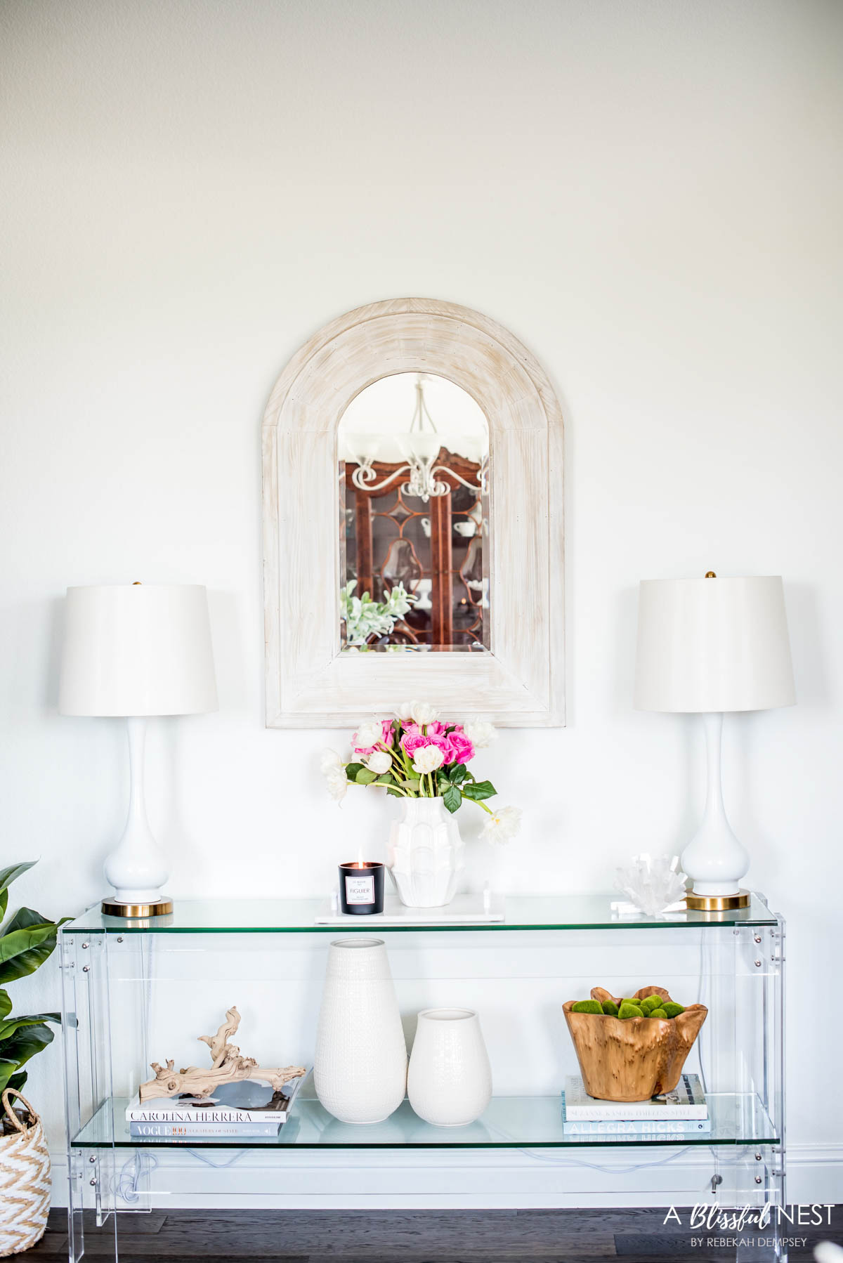 acrylic console table with 2 white table lamps, decorating books stacked, white vases, and a wood bowl filled with moss balls.