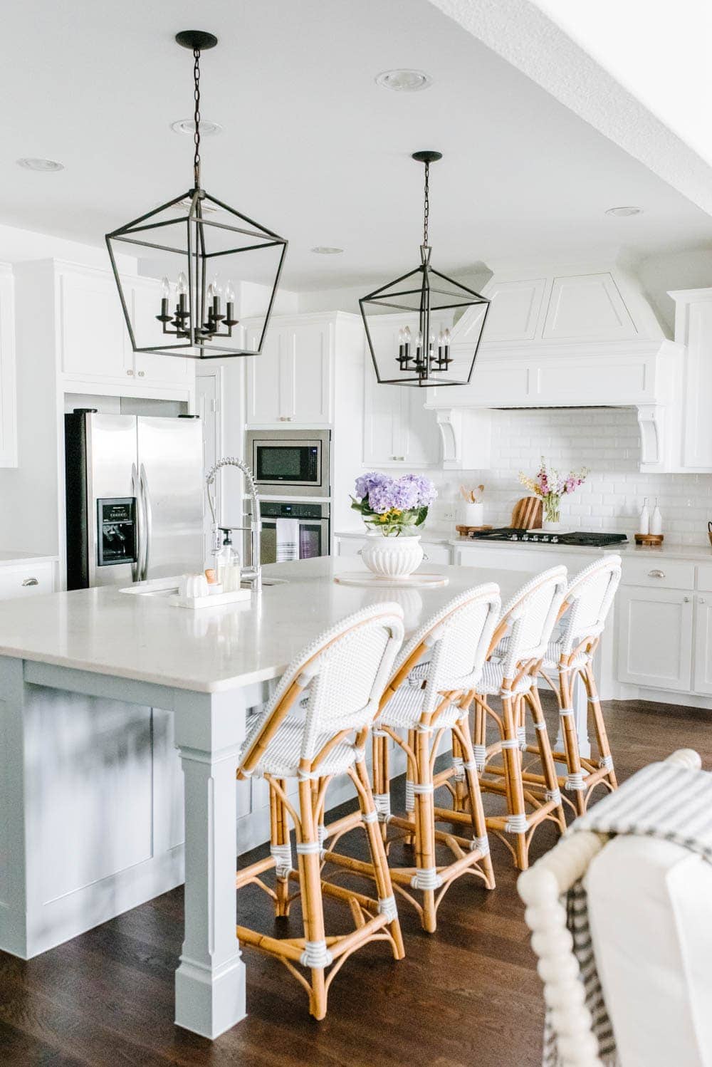 White kitchen cabinets with pale blue kitchen island, black pendant lanterns, and Serena and Lily rattan barstools.