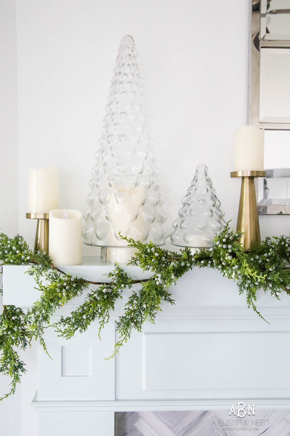 Gorgeous blue and silver Christmas mantle décor with juniper berry garland and glass hurricane Christmas trees. Simple and festive holiday décor in this open concept living space. Check out all the white, silver and gold Christmas decor in this holiday home tour on ABlissfulNest.com. #ABlissfulNest #Christmasdecor #Christmasdecorating #CoastalChristmasdecor #christmastree #christmasmantle