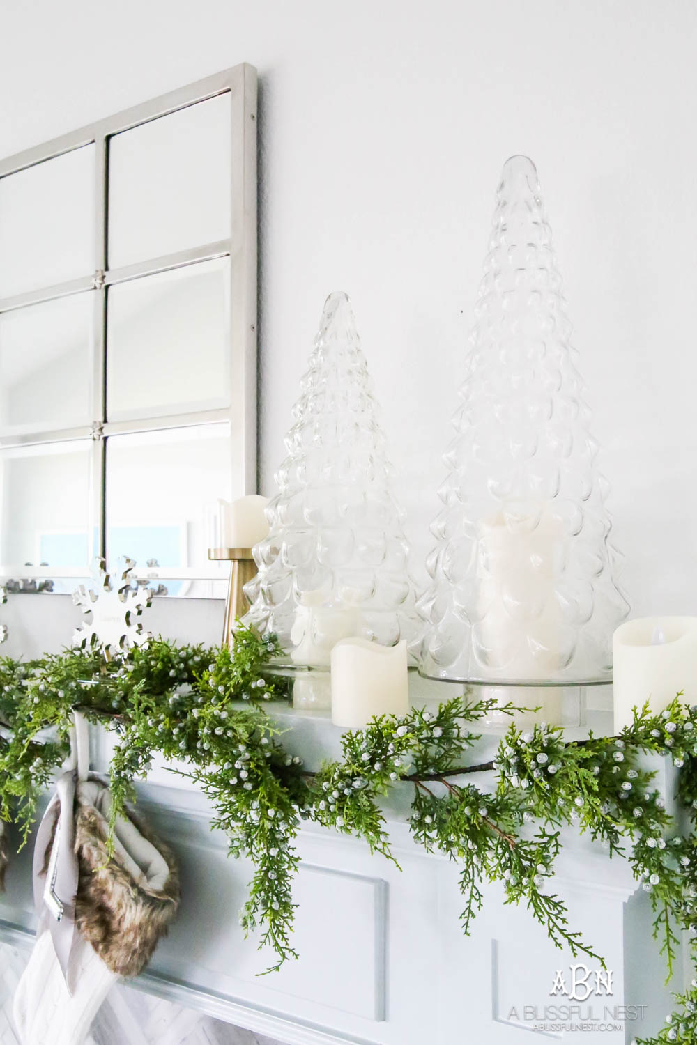 Gorgeous blue and silver Christmas mantle décor with juniper berry garland and glass hurricane Christmas trees. Simple and festive holiday décor in this open concept living space. Check out all the white, silver and gold Christmas decor in this holiday home tour on ABlissfulNest.com. #ABlissfulNest #Christmasdecor #Christmasdecorating #CoastalChristmasdecor #christmastree #christmasmantle