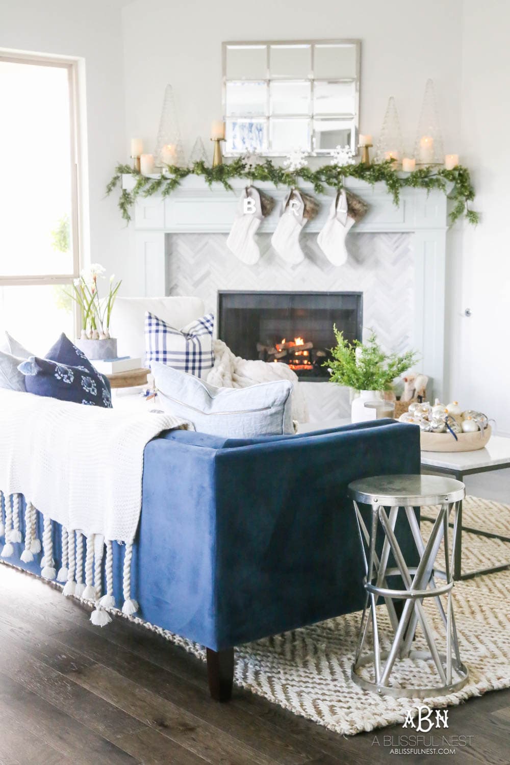 Gorgeous blue and silver Christmas mantle décor with juniper berry garland and glass hurricane Christmas trees. Simple and festive holiday décor in this open concept living space. Check out all the white, silver and gold Christmas decor in this holiday home tour on ABlissfulNest.com. #ABlissfulNest #Christmasdecor #Christmasdecorating #CoastalChristmasdecor #christmastree #christmasmantle