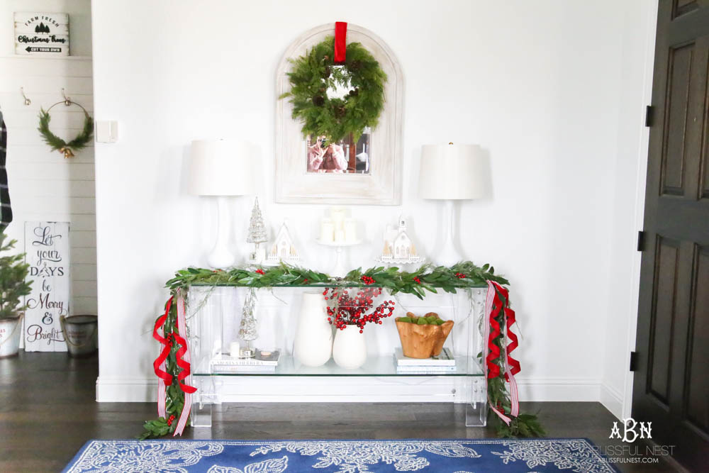 Dress up the entry with classic Christmas décor like a beautiful garland and red and white ribbon. Layered garland creates a fuller look. Add in berries to a vase. Create a festive holiday look in this narrow entryway space. #christmasentryway #christmasentry #christmasentrydecor #christmasentrywayideas #christmashometour