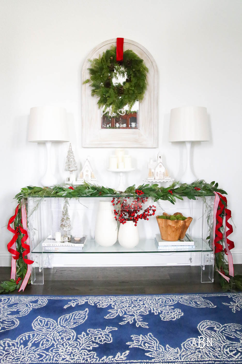 Dress up the entry with classic Christmas décor like a beautiful garland and red and white ribbon. Layered garland creates a fuller look. Add in berries to a vase. Create a festive holiday look in this narrow entryway space. #christmasentryway #christmasentry #christmasentrydecor #christmasentrywayideas #christmashometour