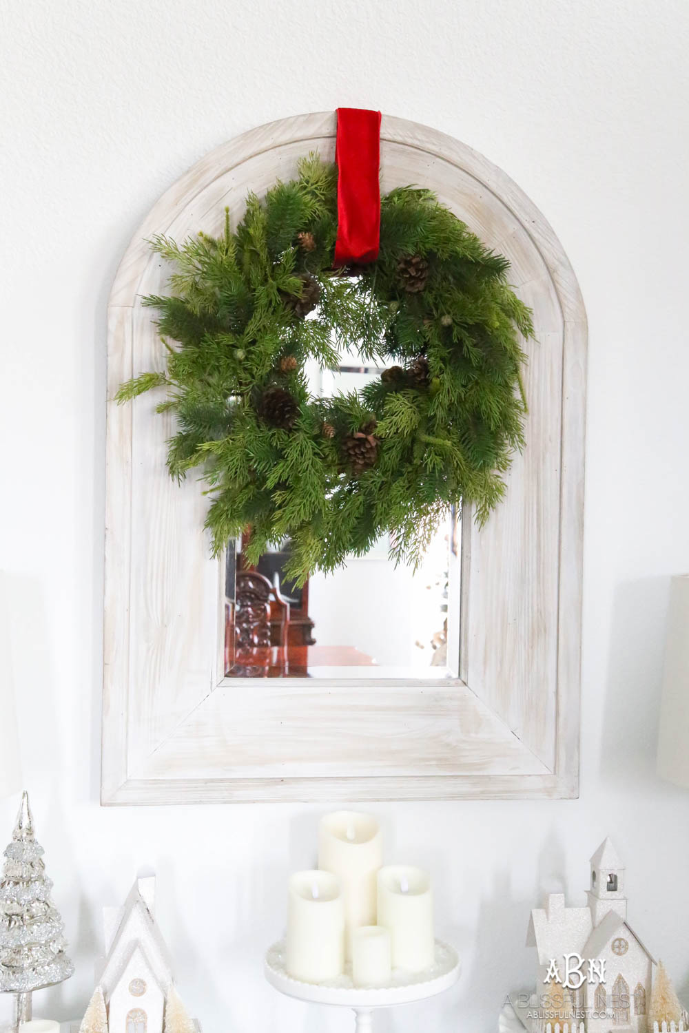 Simple classic Christmas wreath detail on a mirror in an entry. Simple holiday decor that makes a big impact. Red ribbon on a plain wreath. White walls in this holiday home which makes the décor really pop. #christmasentryway #christmasentry #christmasentrydecor #christmasentrywayideas #christmashometour