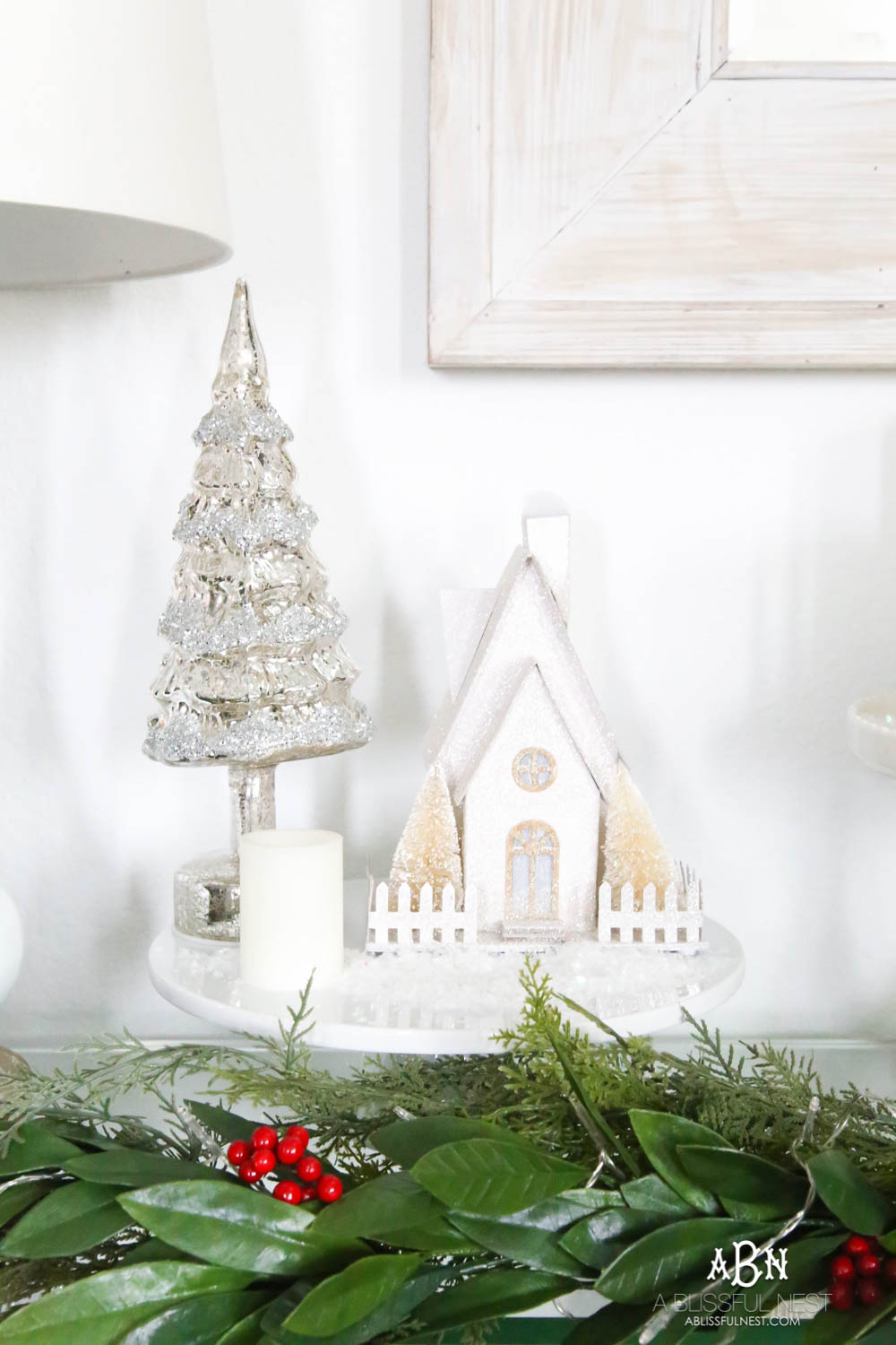 Use cake plates to add height to your display. I love the collection of candles and lighted glitter holiday houses sprinkled with snow for a beautiful simple classic Christmas look. #christmasentryway #christmasentry #christmasentrydecor #christmasentrywayideas #christmashometour