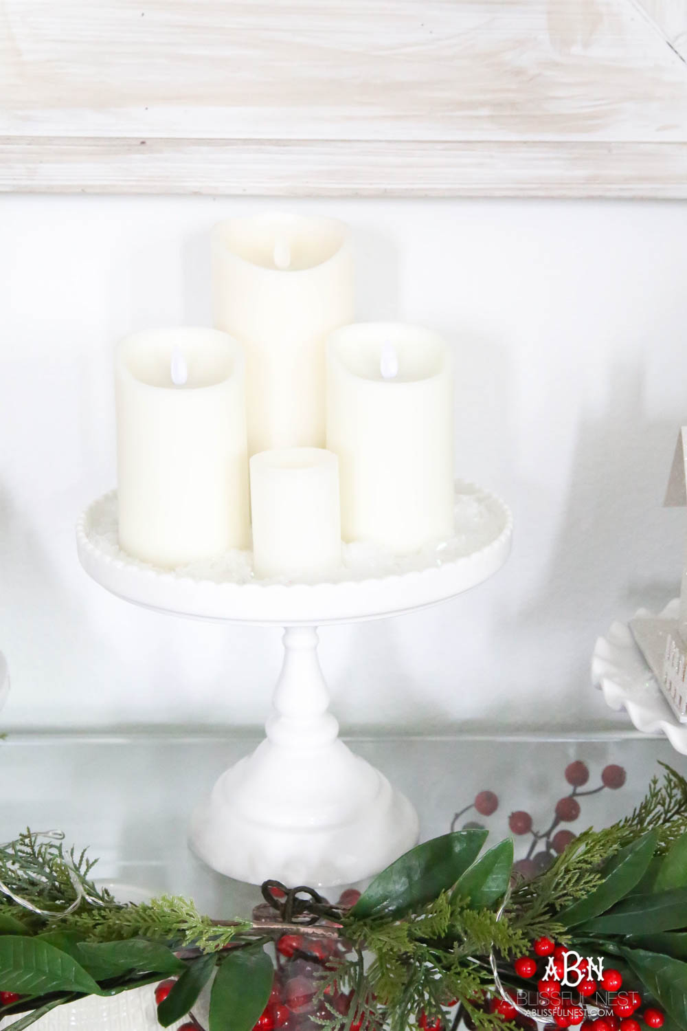 Use cake plates to add height to your display. I love the collection of candles and lighted glitter holiday houses sprinkled with snow for a beautiful simple classic Christmas look. #christmasentryway #christmasentry #christmasentrydecor #christmasentrywayideas #christmashometour