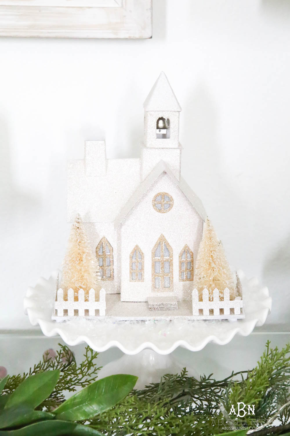 Use cake plates to add height to your display. I love the collection of candles and lighted glitter holiday houses sprinkled with snow for a beautiful simple classic Christmas look. #christmasentryway #christmasentry #christmasentrydecor #christmasentrywayideas #christmashometour