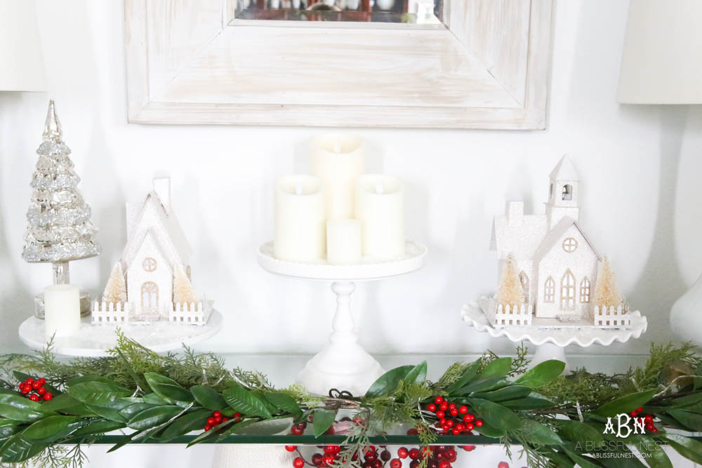 Use cake plates to add height to your display. I love the collection of candles and lighted glitter holiday houses sprinkled with snow for a beautiful simple classic Christmas look. #christmasentryway #christmasentry #christmasentrydecor #christmasentrywayideas #christmashometour