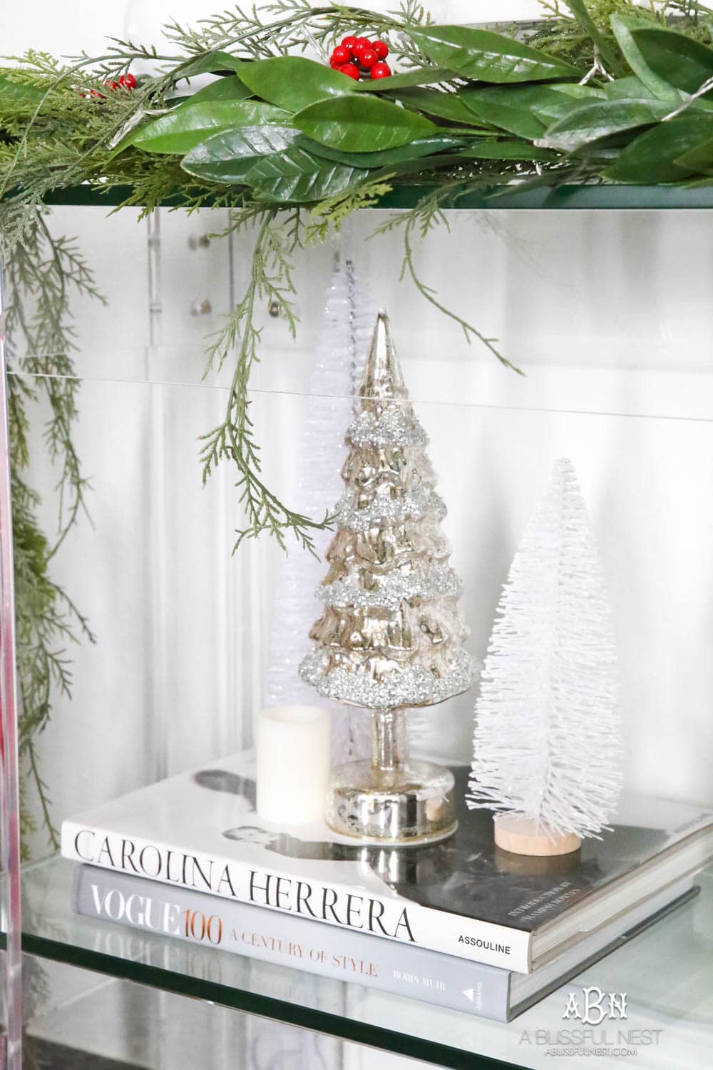 Create a display with your mini trees by clustering them on top of décor books and adding in candles. More ways to add in classic Christmas décor into your home on ablissfulnest.com. #christmasentryway #christmasentry #christmasentrydecor #christmasentrywayideas #christmashometour