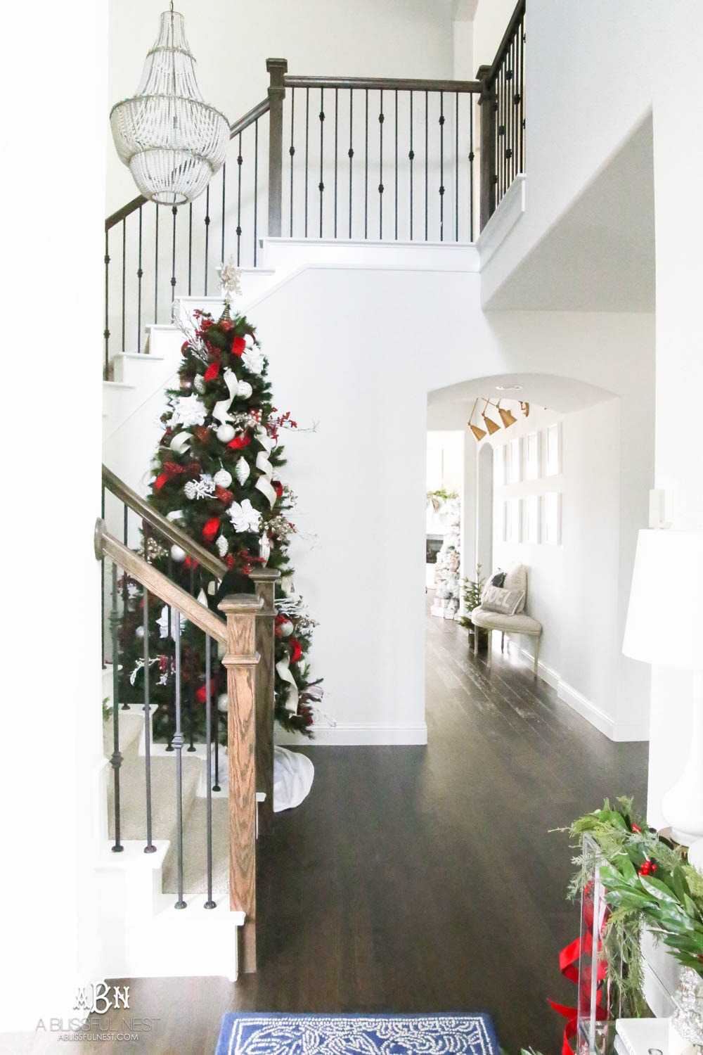 Simple classic Christmas tree and holiday entryway with red and white holiday décor. Christmas tree in stairway with all white walls. #christmasentryway #christmasentry #christmasentrydecor #christmasentrywayideas #christmashometour