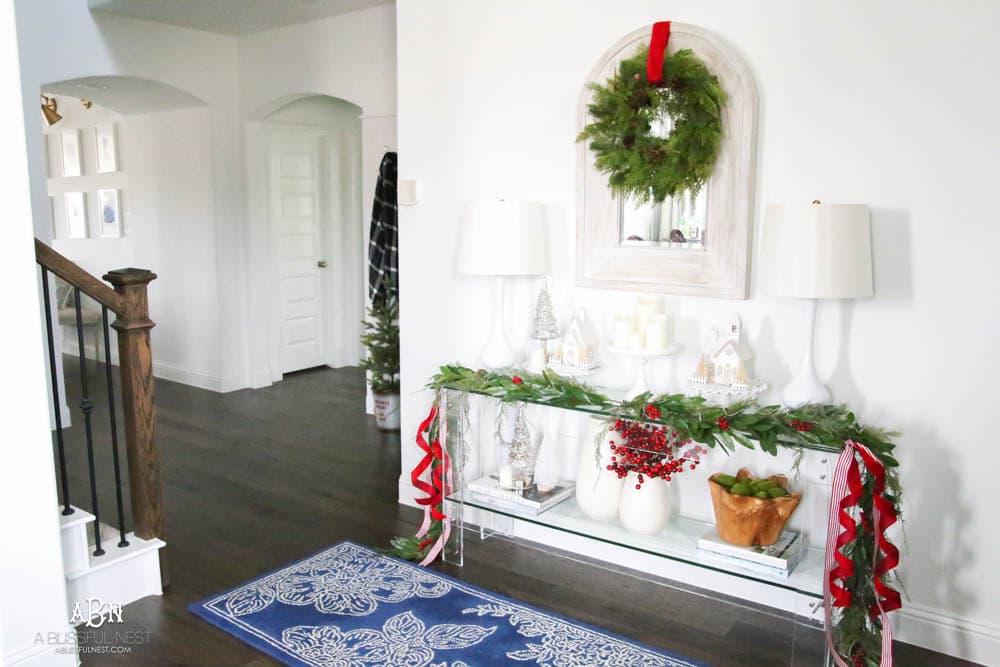 Dress up the entry with classic Christmas décor like a beautiful garland and red and white ribbon. Layered garland creates a fuller look. Add in berries to a vase. Create a festive holiday look in this narrow entryway space. #christmasentryway #christmasentry #christmasentrydecor #christmasentrywayideas #christmashometour