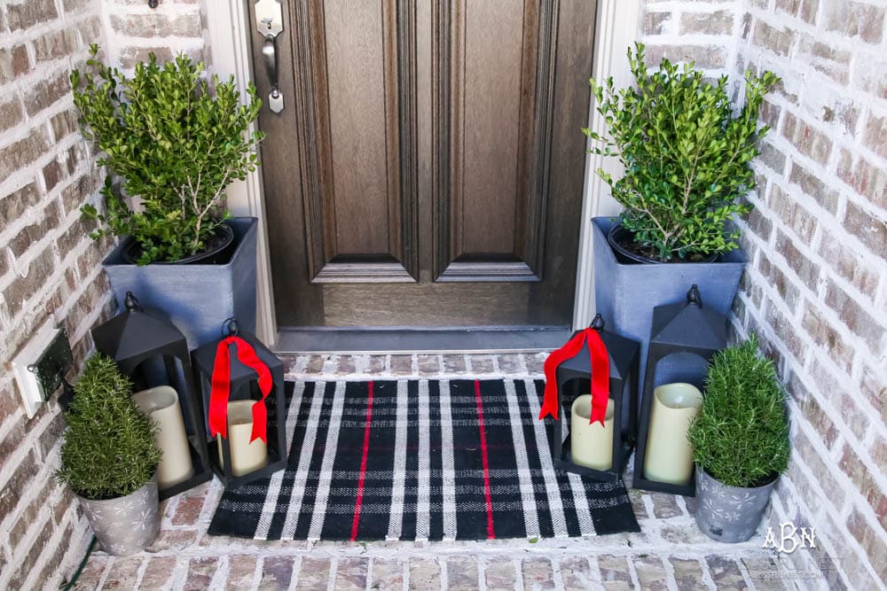 Classic Christmas front porch with red, white and black details. Festive plaid door mat and simple black lanterns. Red Christmas balls on a beautiful Christmas wreath. #christmasporch #christmasfrontporch #christmasporchdecor #christmasporchideas #christmashometou