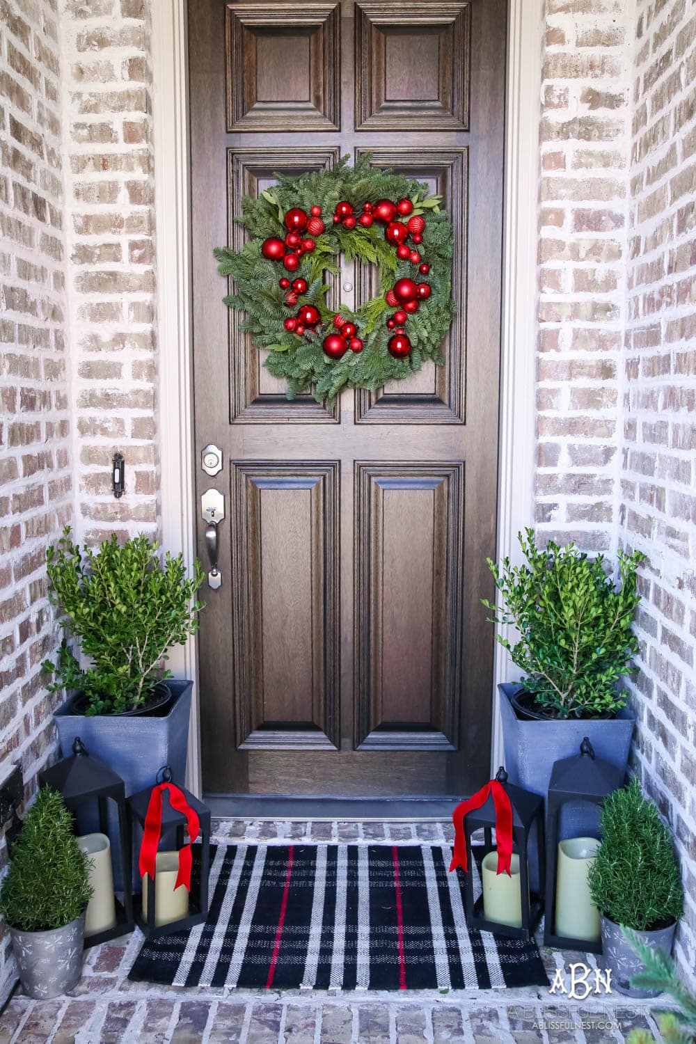 Classic Christmas front porch with red, white and black details. Festive plaid door mat and simple black lanterns. Red Christmas balls on a beautiful Christmas wreath. #christmasporch #christmasfrontporch #christmasporchdecor #christmasporchideas #christmashometour