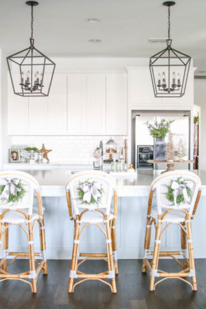 Blue and silver Christmas kitchen décor. All white kitchen with subtle holiday décor for a fresh coastal Christmas look. Mini wreaths on the backs of the barstools, beautiful bay leaf above the range. #christmaskitchen #christmashometour #holidayhometour #kitchenideas