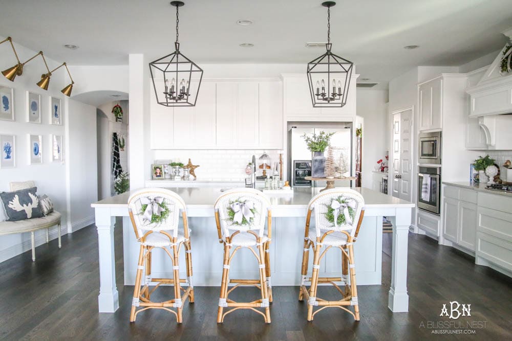 Blue and silver Christmas kitchen décor. All white kitchen with subtle holiday décor for a fresh coastal Christmas look. Mini wreaths on the backs of the barstools, beautiful bay leaf above the range. #christmaskitchen #christmashometour #holidayhometour #kitchenideas