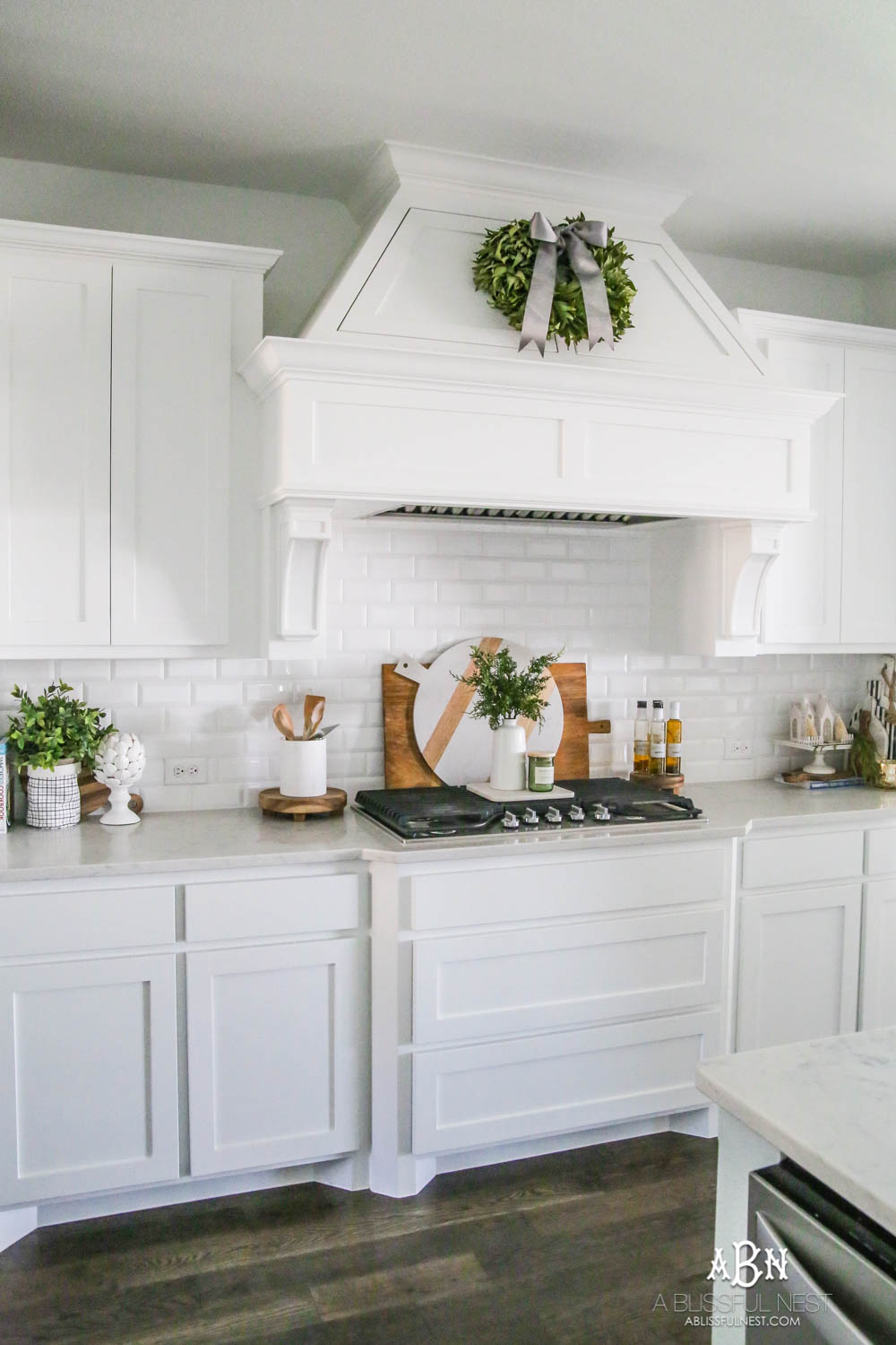 Blue and silver Christmas kitchen décor. All white kitchen with subtle holiday décor for a fresh coastal Christmas look. Mini wreaths on the backs of the barstools, beautiful bay leaf above the range. #christmaskitchen #christmashometour #holidayhometour #kitchenideas