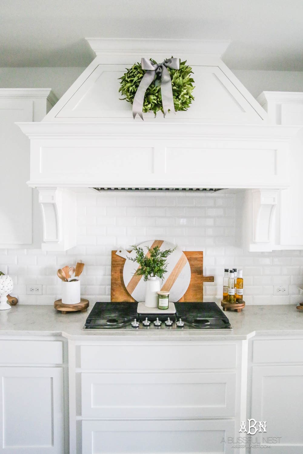 Blue and silver Christmas kitchen décor. All white kitchen with subtle holiday décor for a fresh coastal Christmas look. Mini wreaths on the backs of the barstools, beautiful bay leaf above the range. #christmaskitchen #christmashometour #holidayhometour #kitchenideas