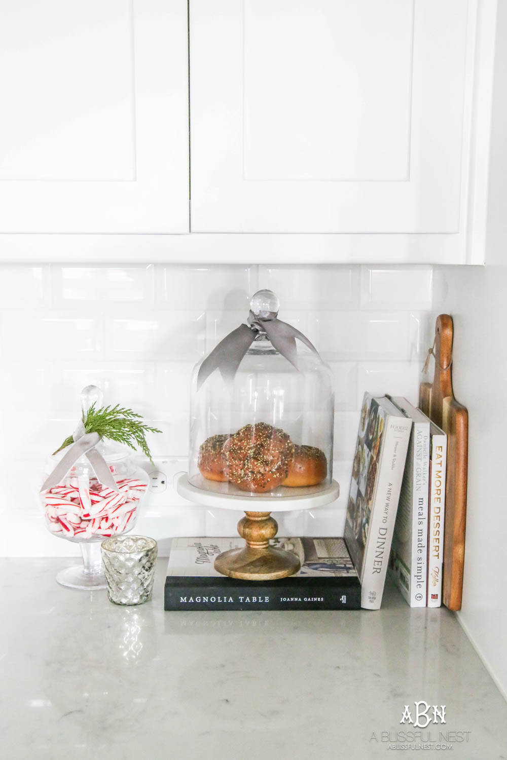 Blue and silver Christmas kitchen décor. All white kitchen with subtle holiday décor for a fresh coastal Christmas look. Mini wreaths on the backs of the barstools, beautiful bay leaf above the range. #christmaskitchen #christmashometour #holidayhometour #kitchenideas