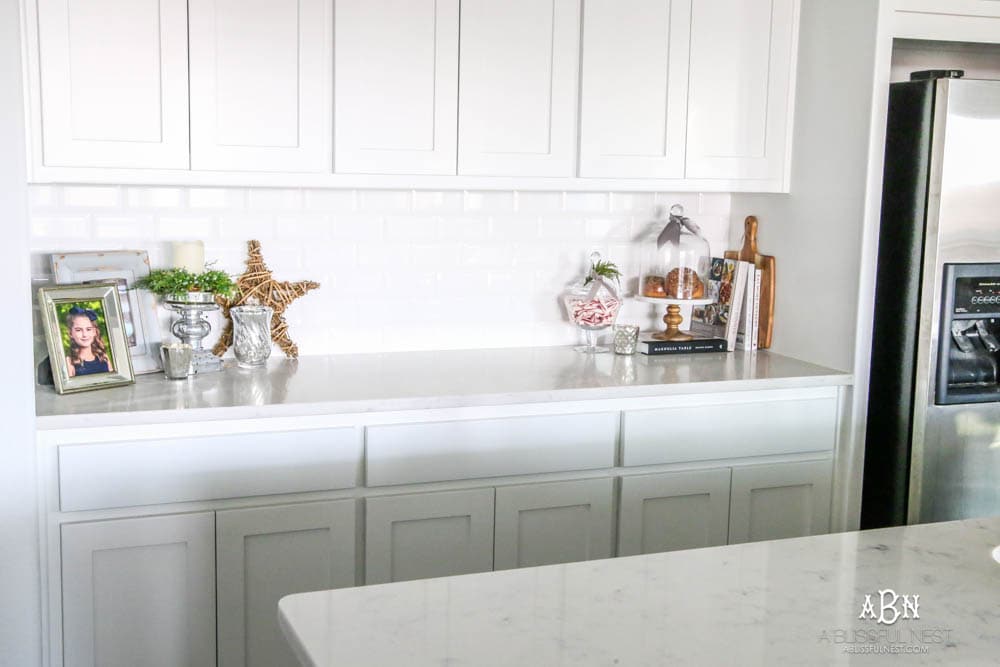 Blue and silver Christmas kitchen décor. All white kitchen with subtle holiday décor for a fresh coastal Christmas look. Mini wreaths on the backs of the barstools, beautiful bay leaf above the range. #christmaskitchen #christmashometour #holidayhometour #kitchenideas