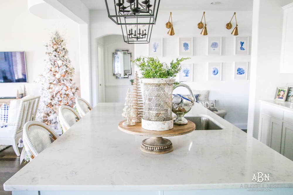 Blue and silver Christmas kitchen décor. All white kitchen with subtle holiday décor for a fresh coastal Christmas look. Mini wreaths on the backs of the barstools, beautiful bay leaf above the range. #christmaskitchen #christmashometour #holidayhometour #kitchenideas