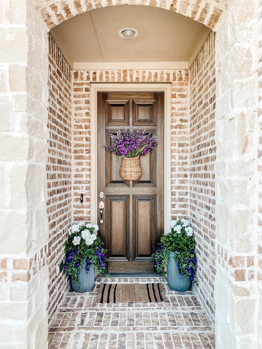 A front porch with purple artificial flowers. 2 planters on either side of the door with white geraniums.