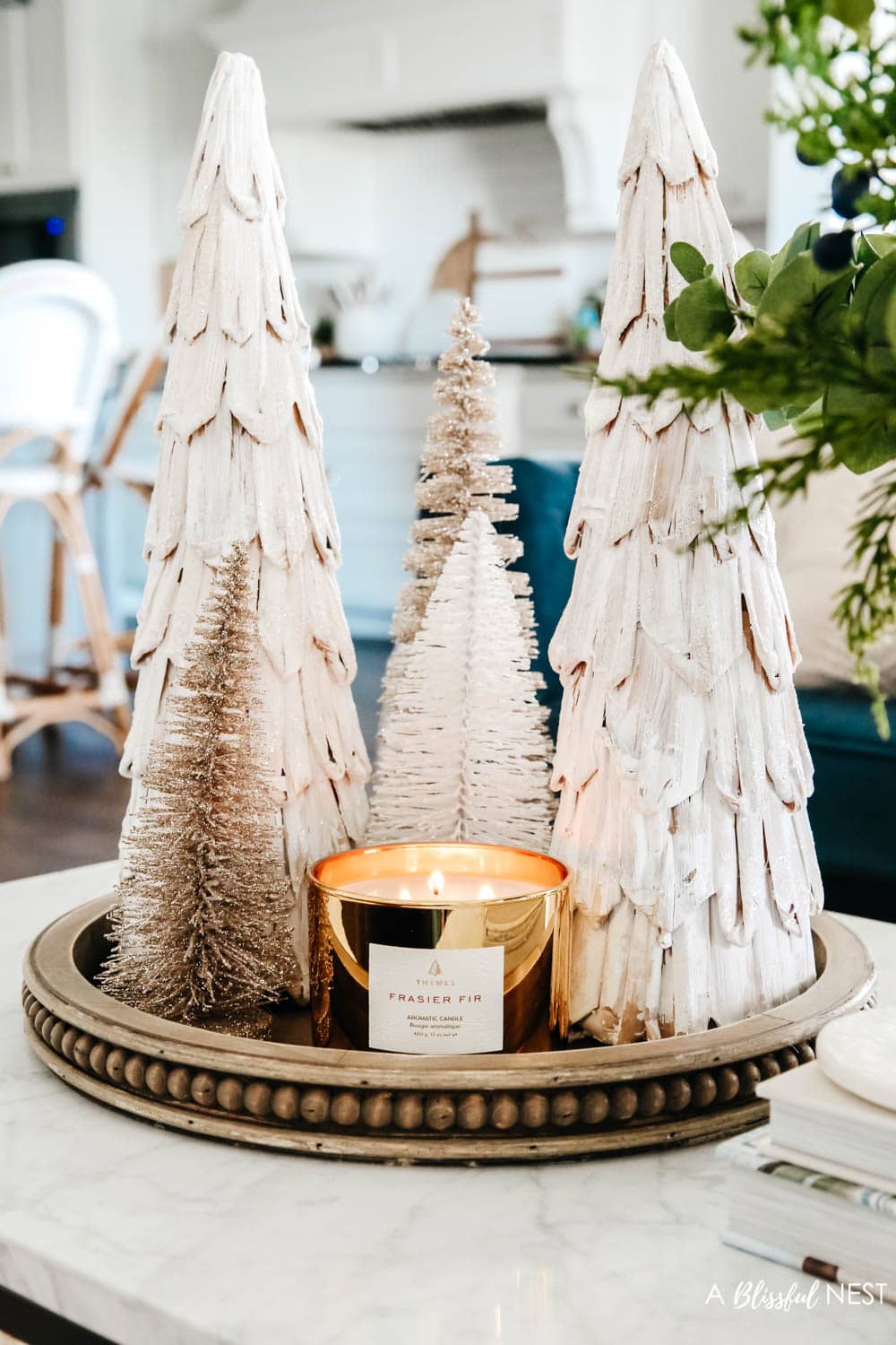 Cluster of mini Christmas trees on wood tray with a gold candle all on a coffee table in front of a blue sofa.