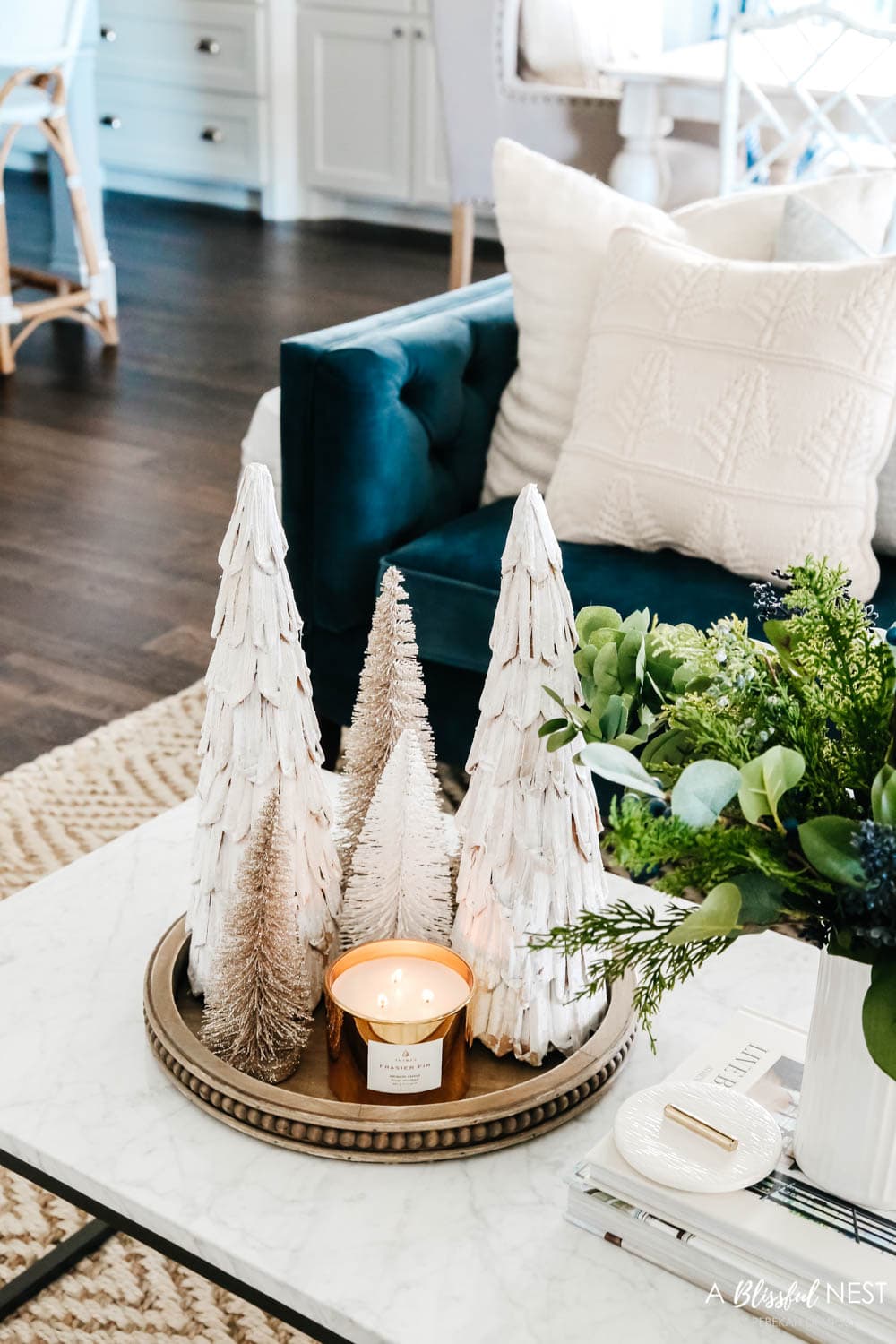 Cluster of mini Christmas trees on wood tray with a gold candle all on a coffee table in front of a blue sofa.