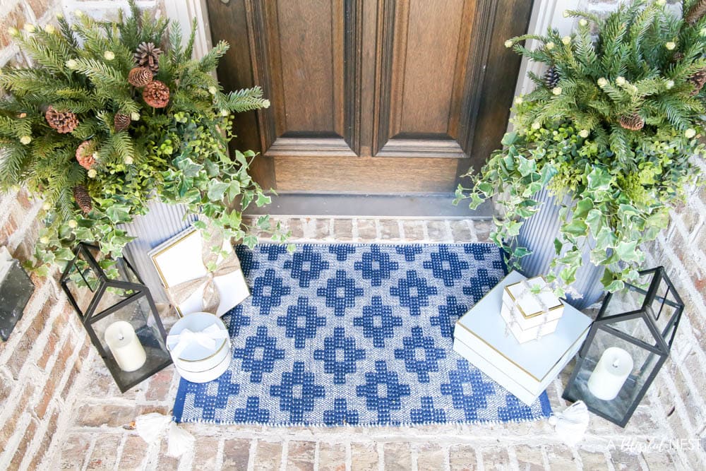 Brick entryway with solid wood door, pine wreath with a white satin ribbon, blue and white doormat with planters and black lanterns with gift boxes tucked next to them.