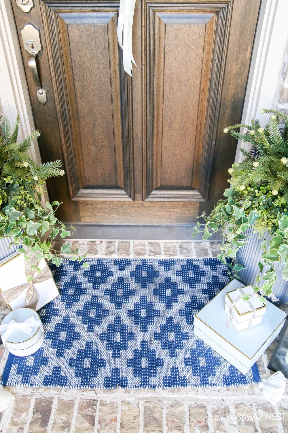 Brick entryway with solid wood door, pine wreath with a white satin ribbon, blue and white doormat with planters and black lanterns with gift boxes tucked next to them.