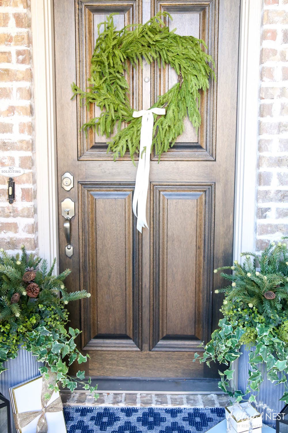 Brick entryway with solid wood door, pine wreath with a white satin ribbon, blue and white doormat with planters and black lanterns with gift boxes tucked next to them.
