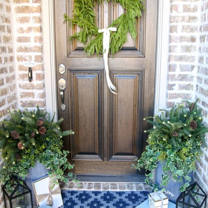 Brick entryway with solid wood door, pine wreath with a white satin ribbon, blue and white doormat with planters and black lanterns with gift boxes tucked next to them.