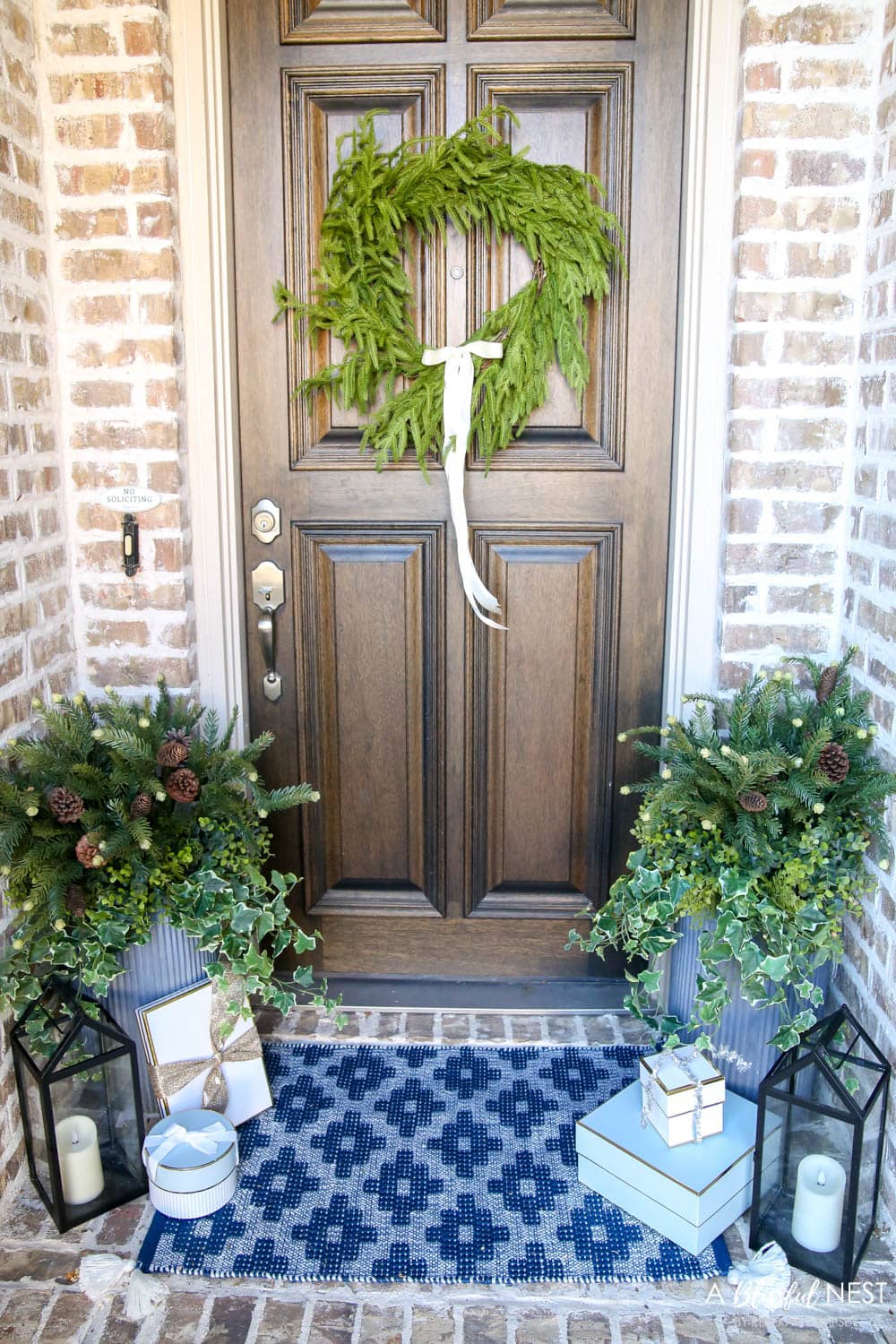 Brick entryway with solid wood door, pine wreath with a white satin ribbon, blue and white doormat with planters and black lanterns with gift boxes tucked next to them.