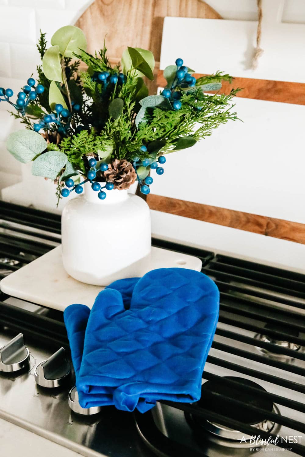 A white vase with greenery and berries, navy potholders sitting on a stove with cutting boards behind it.