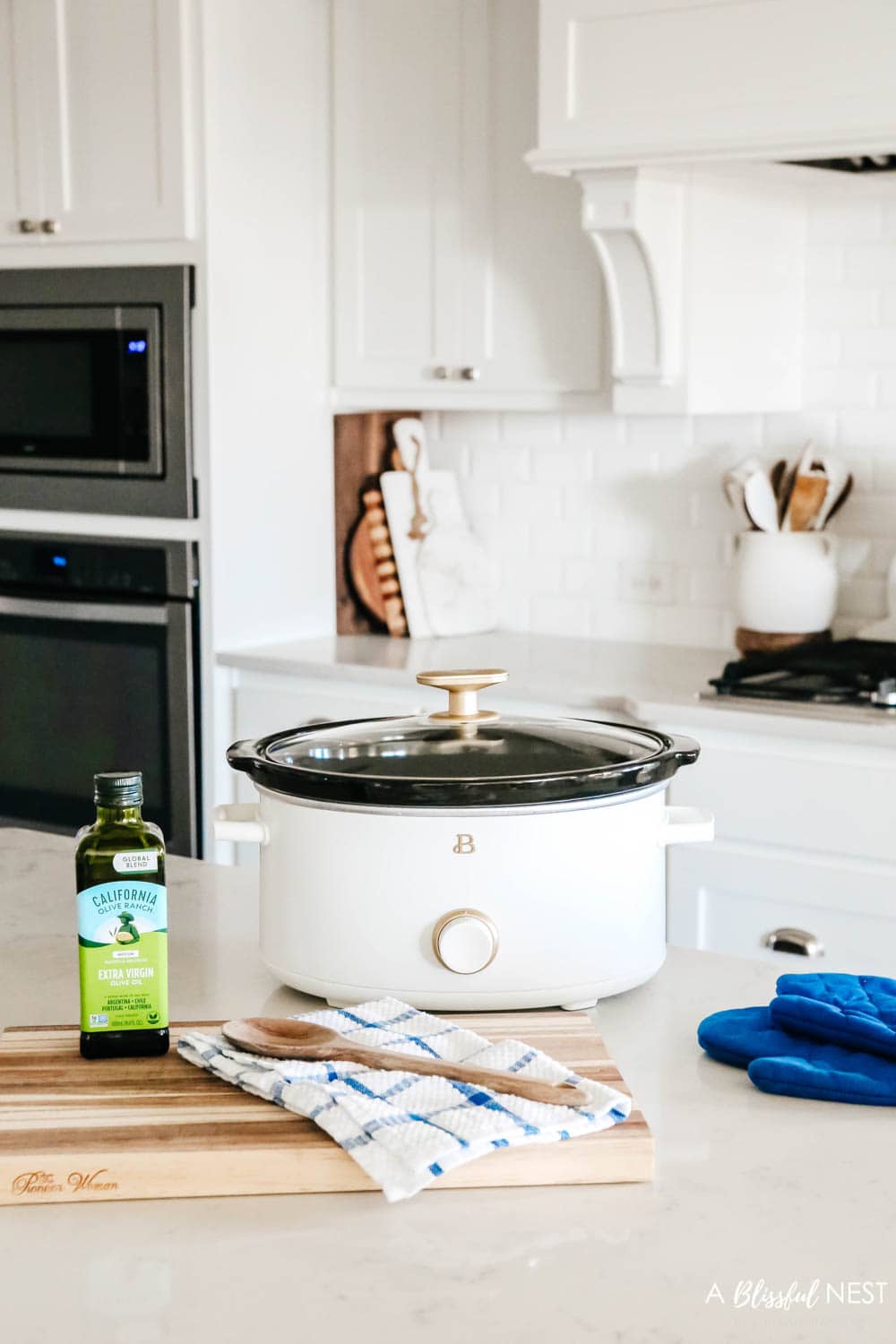 A white kitchen, with rattan barstools, white crockpot, navy blue potholders and a cooktop range with a wreath above. 