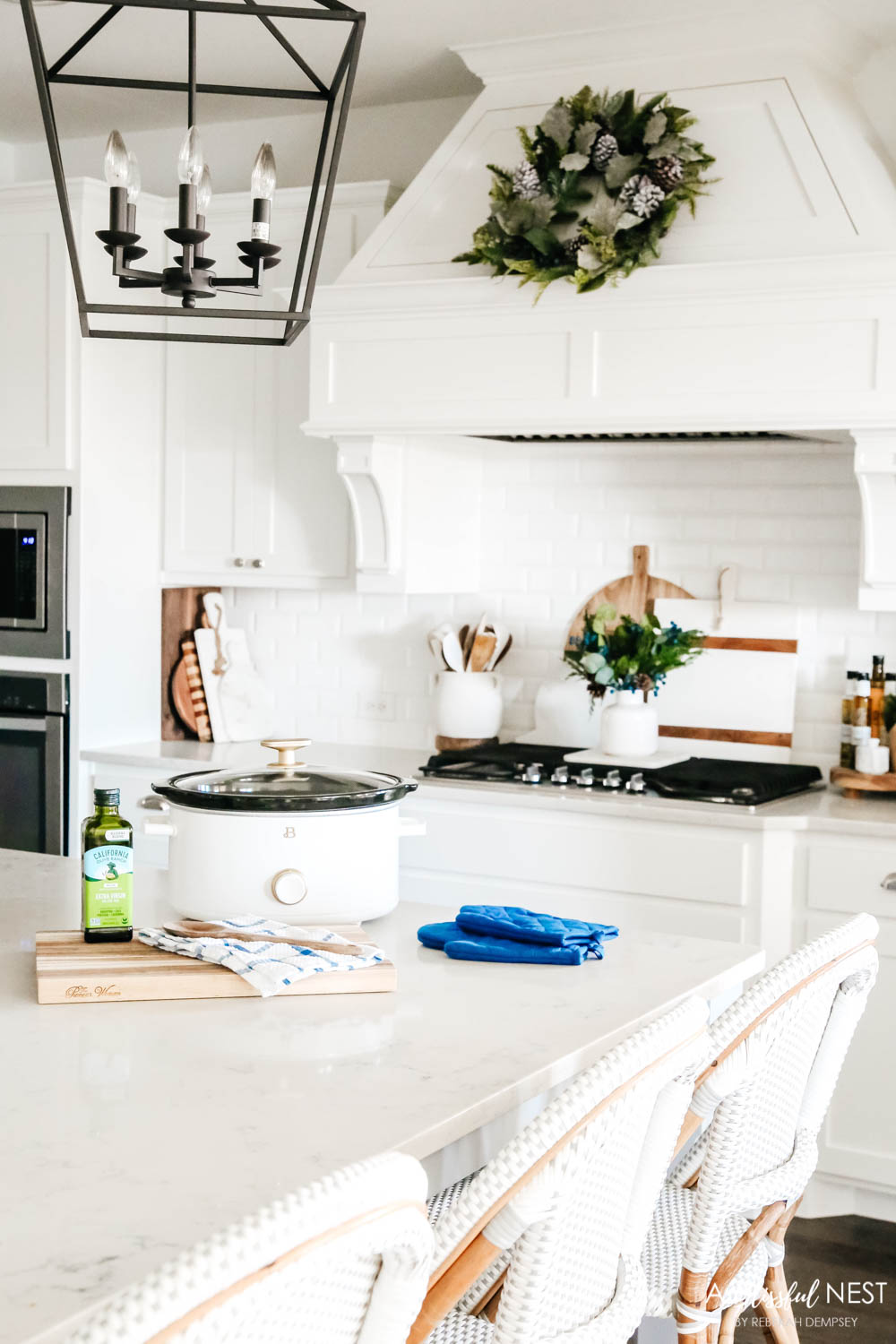 A white kitchen, with rattan barstools, white crockpot, navy blue potholders and a cooktop range with a wreath above.