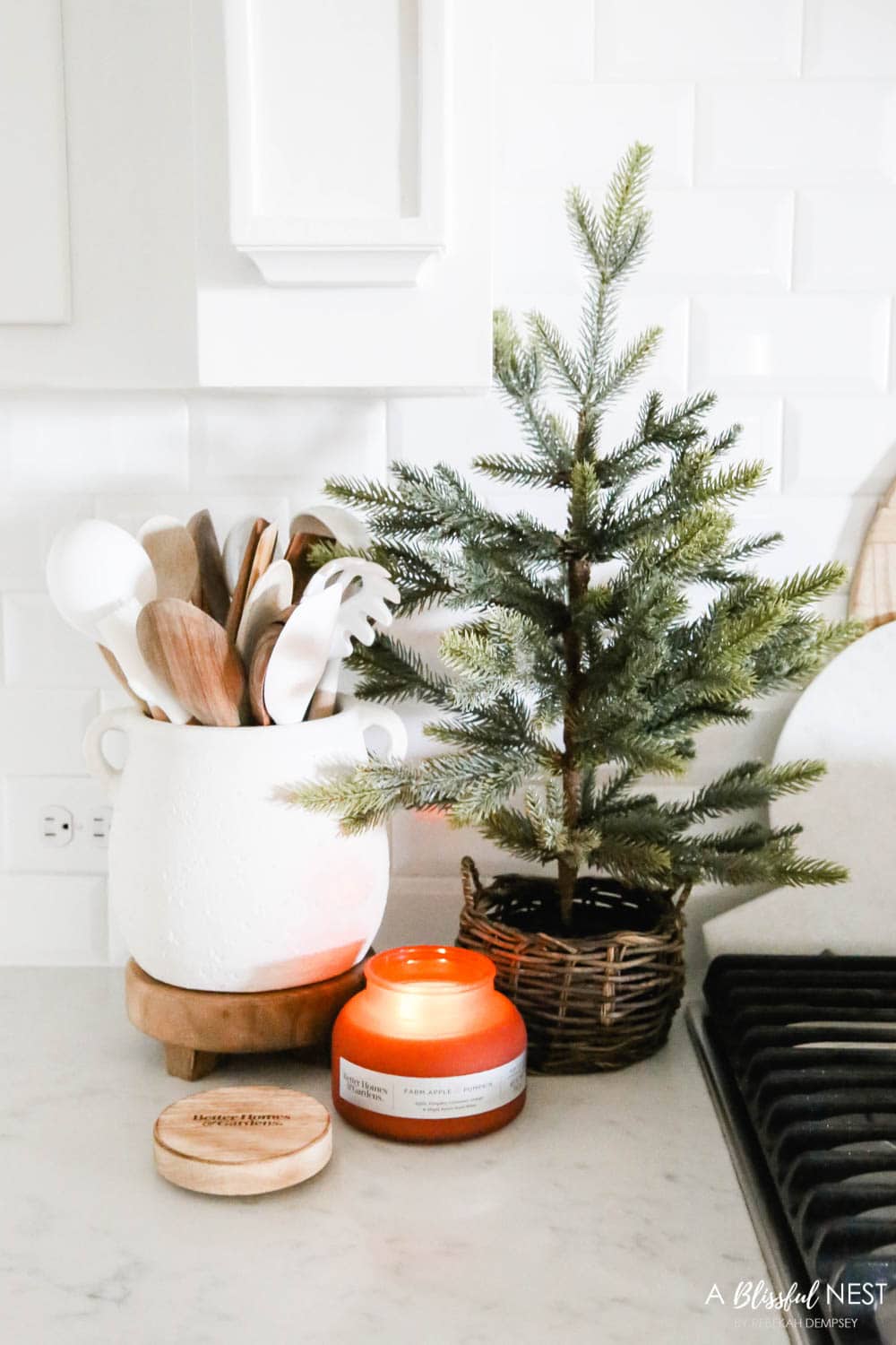 A yummy smelling candle burning in a kitchen next to a cooktop with a cluster of utensils in a white pot and a Christmas tree in a basket.