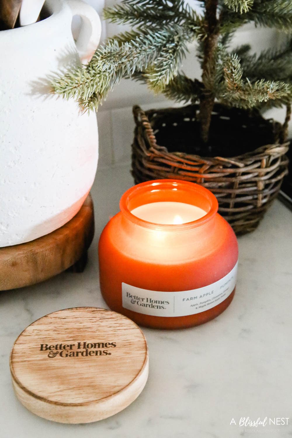 A yummy smelling candle burning in a kitchen next to a cooktop with a cluster of utensils in a white pot and a Christmas tree in a basket.