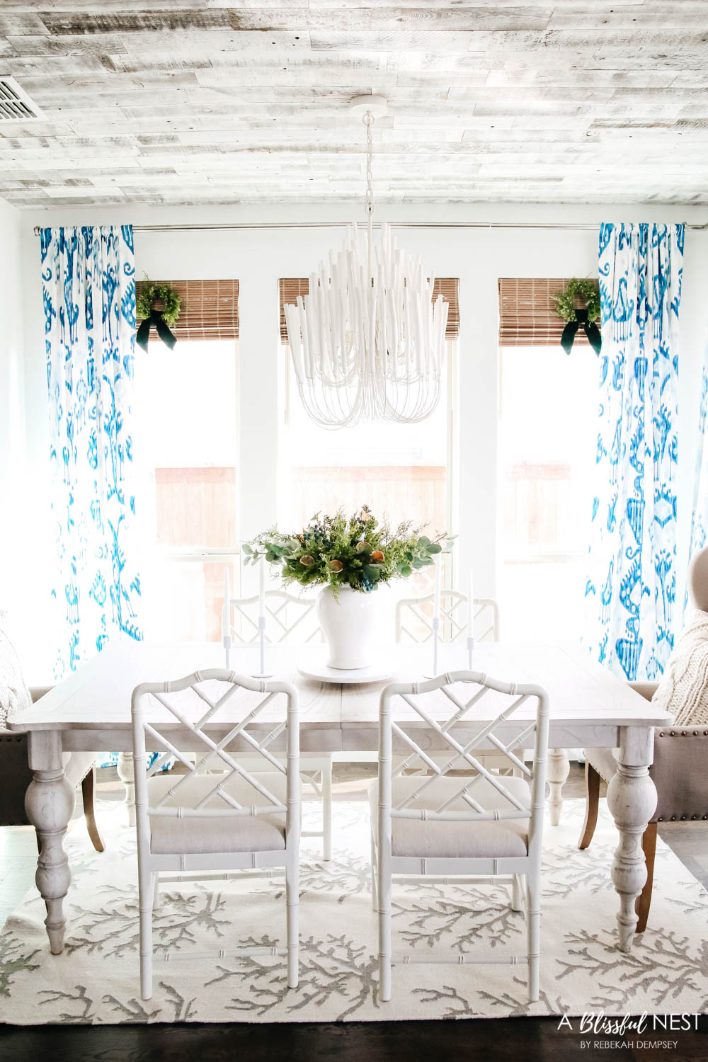 Dining room with white bamboo chairs and blue and white drapes. Simple white candles sticks next to a white vase with juniper floral picks. 
